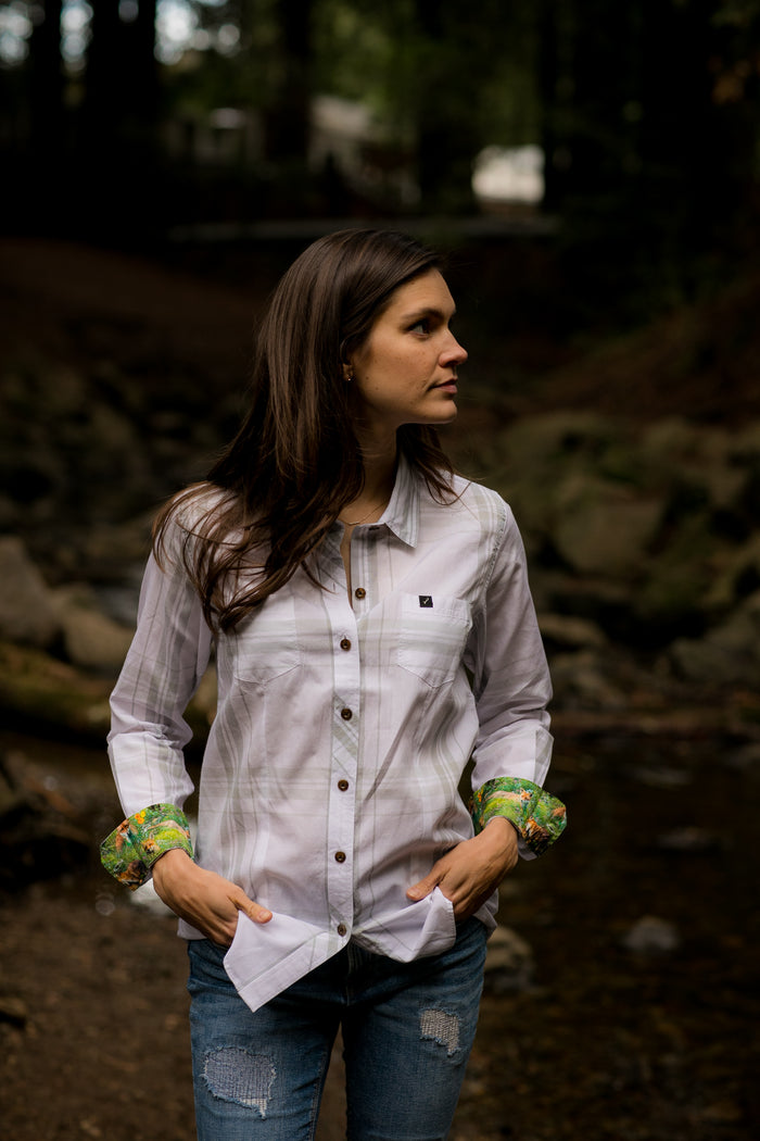 A woman in a Pladra Women's Breeze Shirt- Spring White and jeans stands by a creek, hands in pockets, looking sideways.