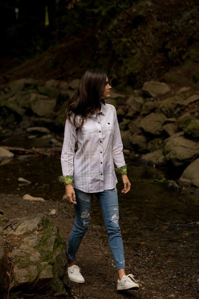 Woman in a Pladra Women's Breeze Shirt - Spring White and jeans stands by a rocky creek in a forest, looking left.
