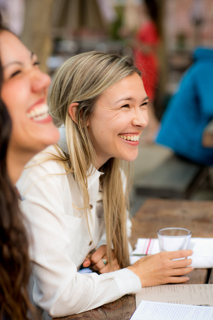 Two women in Pladra Women's Breeze Shirt- Whisper Grey sit outdoors, laughing with papers and a glass of water on the table.