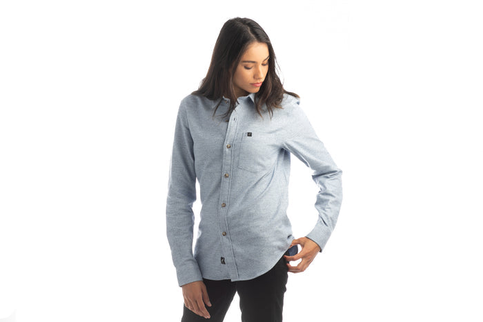 A person with long dark hair, wearing the Women's Cascade Flannel Shirt in Dusty Blue and black pants, stands against a white background, looking down and touching the side of their shirt.