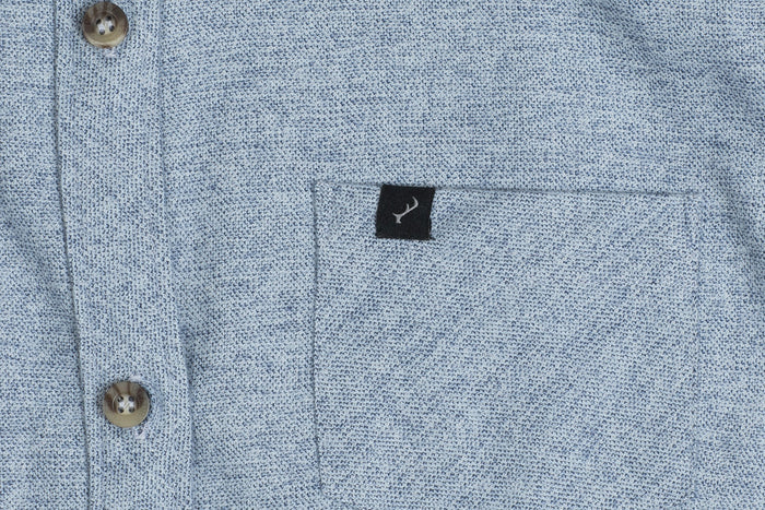 Close-up of the Women's Cascade Flannel Shirt in Dusty Blue, made from heavy Portuguese cotton flannel. Features brown buttons, a front pocket, and a small black tag with a white fish logo on the pocket’s upper edge.