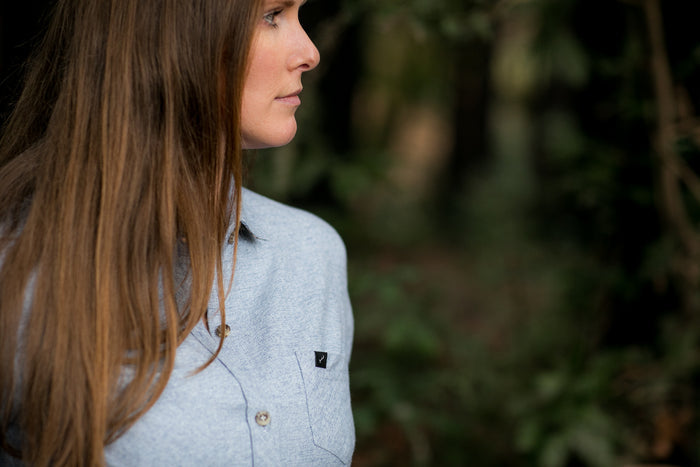 A woman with long brown hair stands outdoors amid trees, wearing the Women's Cascade Flannel Shirt in Dusty Blue, made from heavy brushed cotton.
