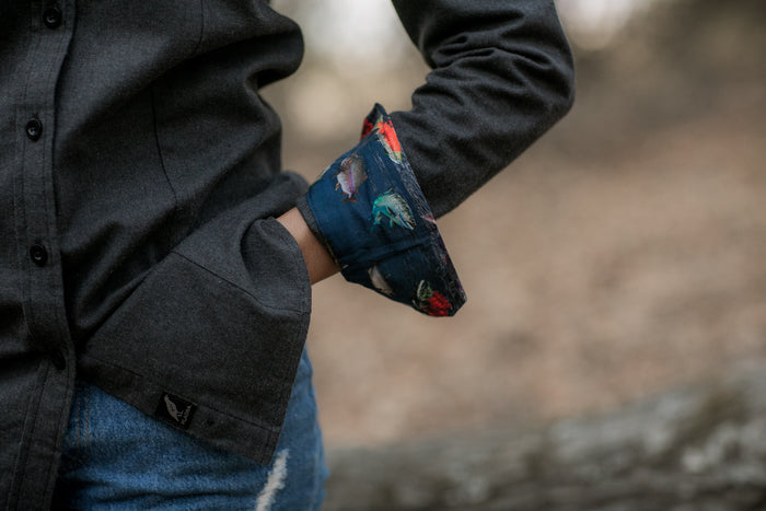 A woman stands outdoors in blue jeans and the Women's Cascade Flannel Shirt in Jet Black Heather, one hand in her pocket. The background is blurred and natural.