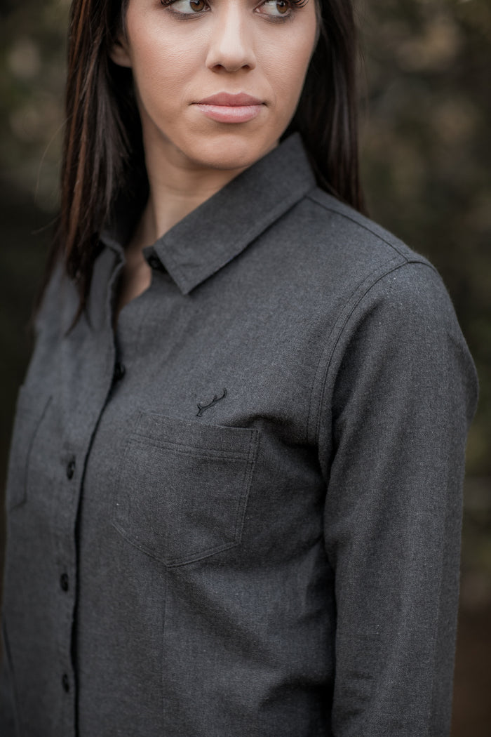 A woman with long dark hair wears the Women's Cascade Flannel Shirt in Jet Black Heather, featuring a chest pocket, as she stands outdoors against a blurred background and looks to the side with a neutral expression.