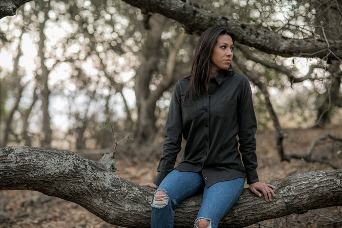 A woman with long dark hair, wearing the Women's Cascade Flannel Shirt - Jet Black Heather and ripped blue jeans, sits on a large tree branch in a wooded area with dry leaves and branches, gazing to her right.
