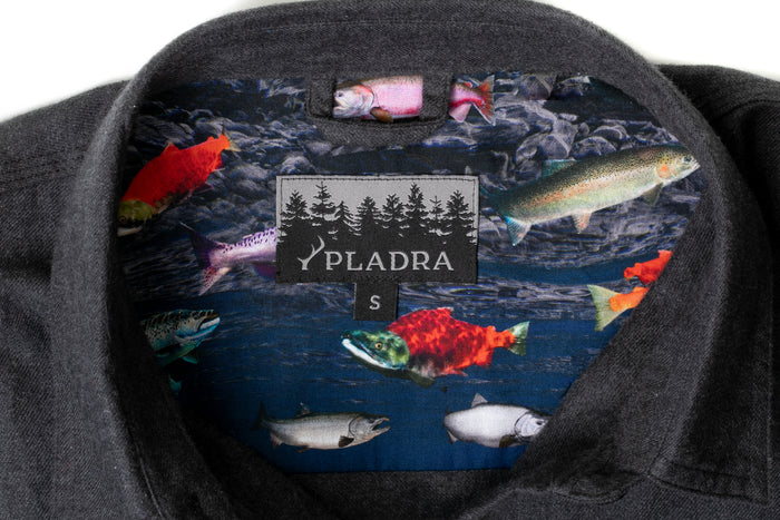 Close-up of the inside collar of the Women's Cascade Flannel Shirt - Jet Black Heather, featuring a colorful fish-patterned lining, PLADRA label, size S tag, and 100% cotton quality.