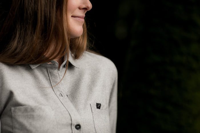A woman with long hair smiles softly, wearing the Pladra Women's Cascade Flannel Shirt in Stratus Grey outdoors.