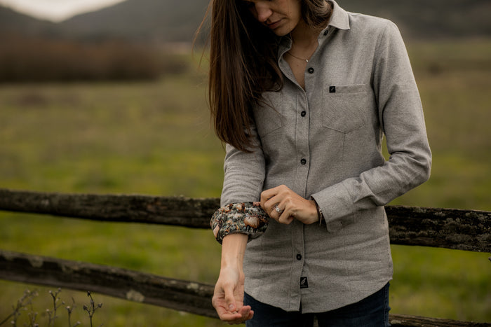 A woman in a Pladra Women's Cascade Flannel Shirt in Stratus Grey rolls up her sleeve by a wooden fence, hills beyond.