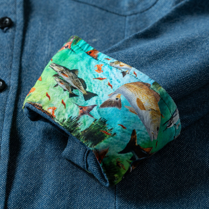 A close-up of the Women's Cascade Shirt in Biscayne Blue Twill shows the cuff turned up, revealing a vibrant inner lining with an underwater fish scene featuring colorful fish among green aquatic plants.