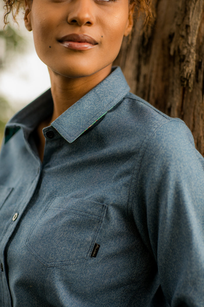 A woman in the Women's Cascade Shirt - Biscayne Blue Twill leans against a tree outdoors. The image, cropped from shoulders up, captures her subtle smile against a natural backdrop, highlighting the shirt's sustainable cotton fabric.