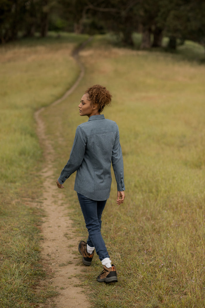 A woman with curly hair, wearing the Women's Cascade Shirt in Biscayne Blue Twill and jeans, strolls a narrow dirt path through a grassy field, glancing back over her shoulder as trees and greenery fill the background.