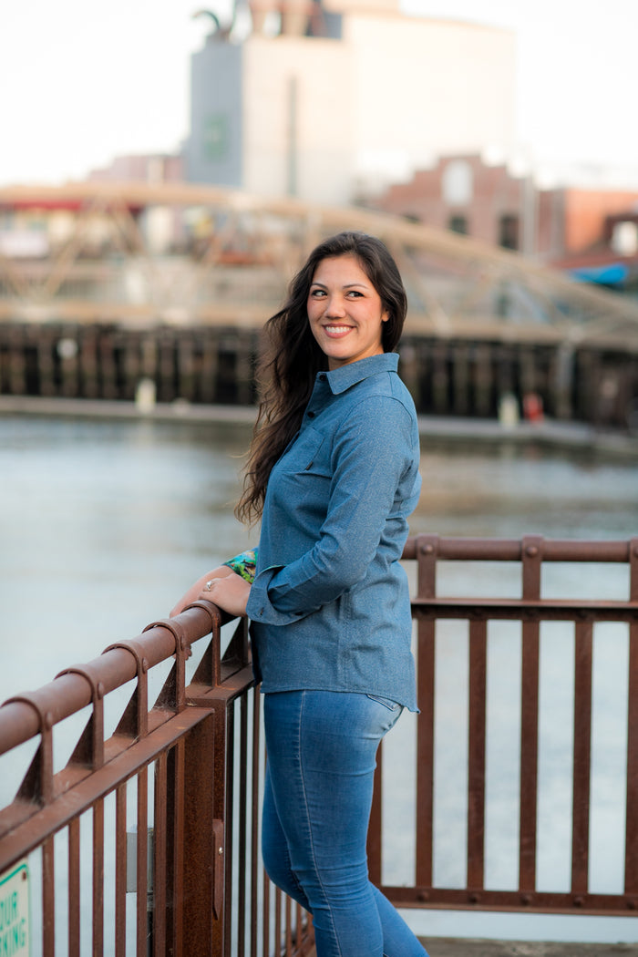 A woman with long dark hair, wearing the Women's Cascade Shirt in Biscayne Blue Twill and jeans, smiles by a brown metal railing overlooking the water, with industrial buildings in the background.
