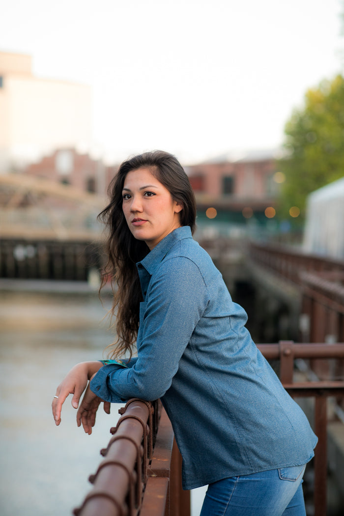A woman with long brown hair, wearing the Women's Cascade Shirt in Biscayne Blue Twill, leans on a metal railing outdoors with blurred buildings and trees in the background.