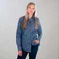 A woman with long blonde hair wears the Women's Cascade Shirt- Indigo Blue Chambray and dark pants, standing against a light gray background with one hand in her pocket and a neutral expression.