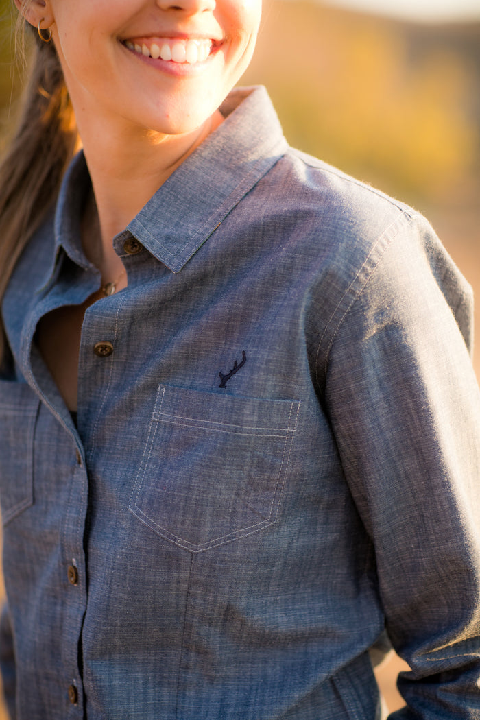 A smiling woman stands outdoors in warm sunlight wearing the Women's Cascade Shirt in Indigo Blue Chambray, featuring a small embroidered antler logo on the chest pocket.