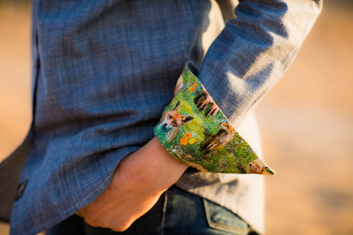A person wears a gray jacket with cuffs turned up to show a fox and green forest print lining. Underneath, they have the Women's Cascade Shirt in Indigo Blue Chambray, made from sustainable cotton, and rest one hand in their pocket.