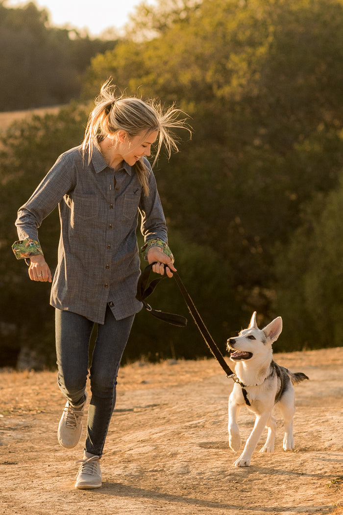 A woman with blonde hair jogging on a dirt path at sunset, smiling at her dog on a leash. She wears the Women's Cascade Shirt in Indigo Blue Chambray, surrounded by green trees under a warm golden glow.