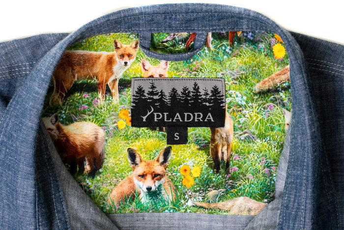 The Women's Cascade Shirt- Indigo Blue Chambray has an inside collar lined with a colorful fox-and-meadow print and a centered black PLADRA S label featuring trees.