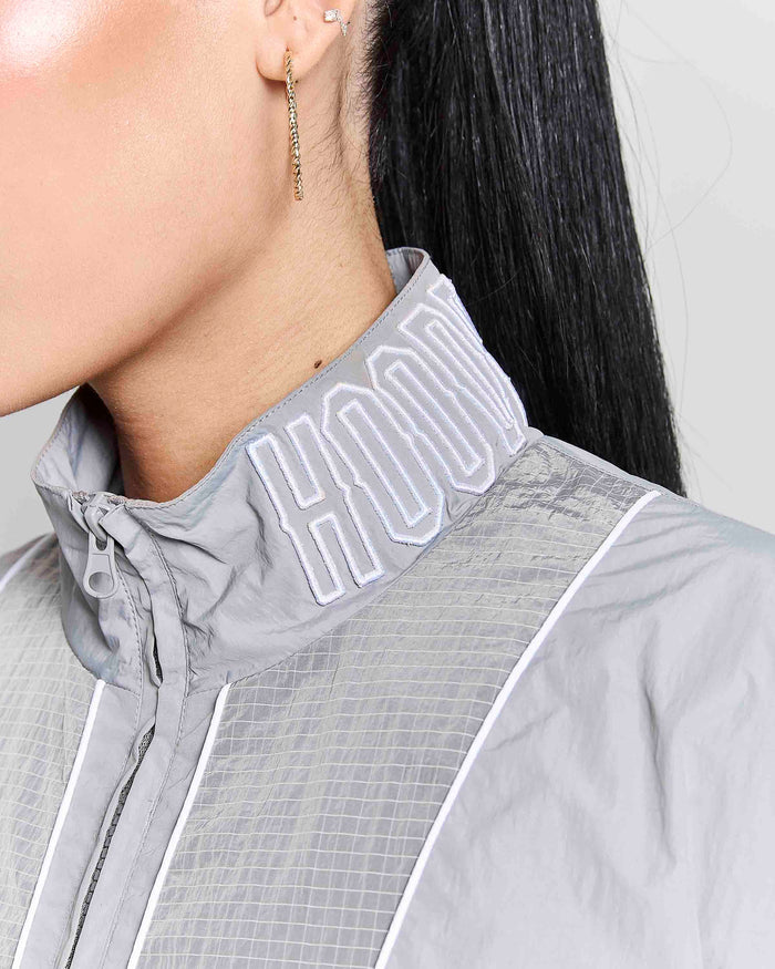 Close-up of a model wearing a light gray athletic jacket with a high collar featuring white embroidered lettering, paired with a gold dangling earring.