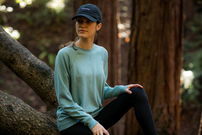 A woman in a Women's Crew Neck Sweatshirt- Bermuda Green Heather sits on a tree branch in a forest, wearing black leggings and a black cap, looking calmly to the side among tall trees and greenery.