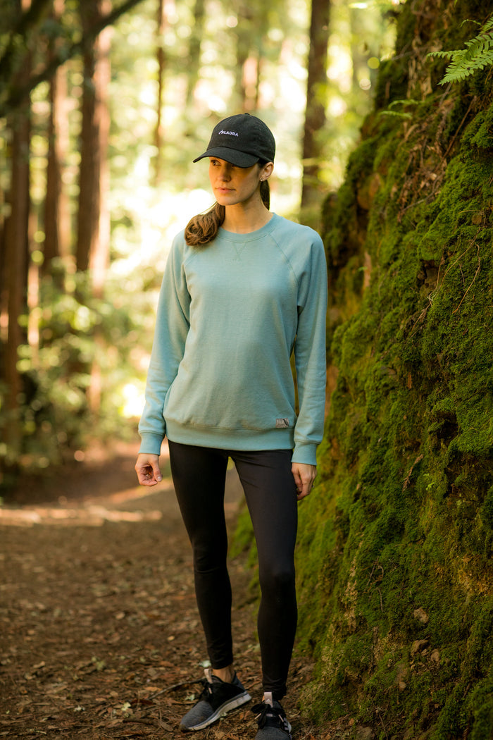 A person in a Bermuda Green Heather Women's Crew Neck Sweatshirt, black leggings, sneakers, and cap stands on a forest trail by a mossy rock, surrounded by tall trees and soft sunlight.