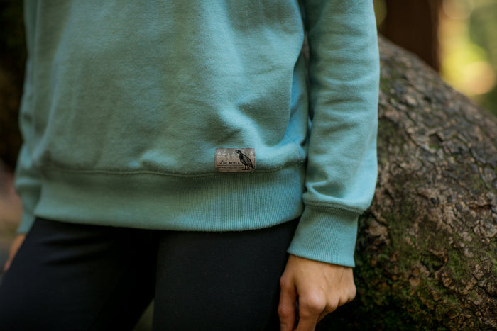 A person wearing a slim fit Women's Crew Neck Sweatshirt in Bermuda Green Heather and black pants stands outdoors by a tree, showing just their torso and arms with a small bird label on the sweatshirt’s hem.