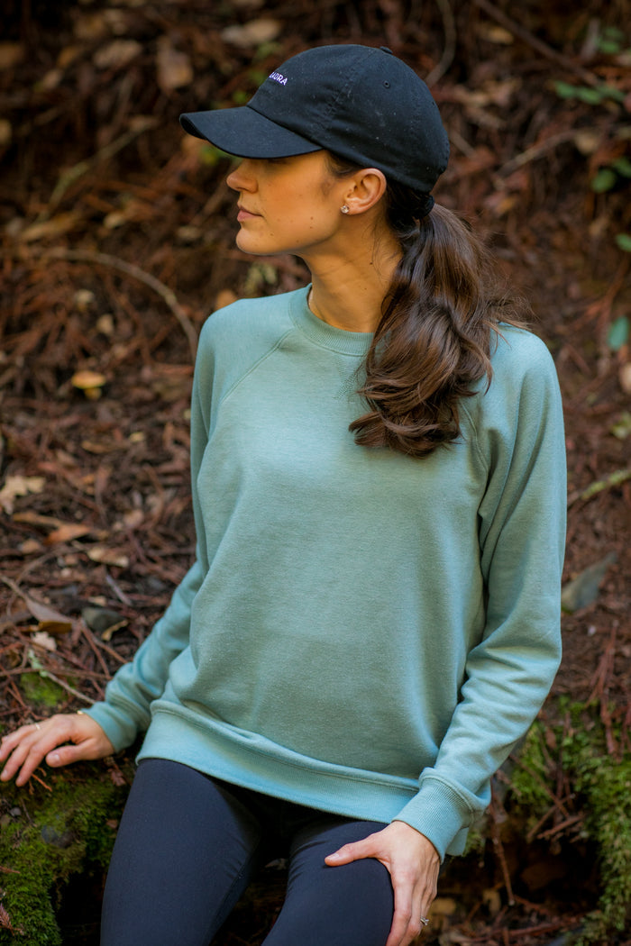 A woman in a slim fit Women's Crew Neck Sweatshirt—Bermuda Green Heather, black cap, and leggings sits outdoors on a mossy rock amid fallen leaves and branches, looking to the side.