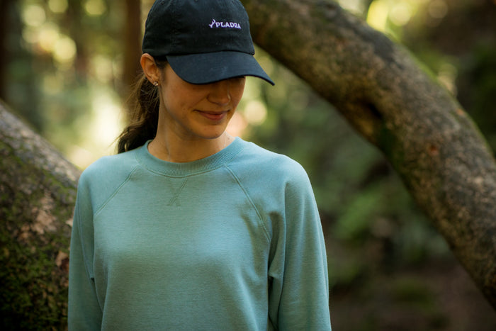 A woman stands outdoors in a forest, sunlight filtering through the trees, wearing a black cap and the Women's Crew Neck Sweatshirt in Bermuda Green Heather. She looks down with a slight smile amidst blurred branches.
