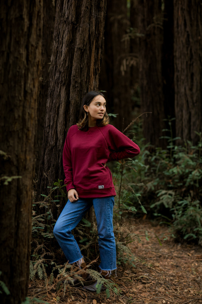 A woman in a Pladra Women's Crew Neck Sweatshirt–Pomegranate Red and jeans stands in a forest, leaning against a tree.