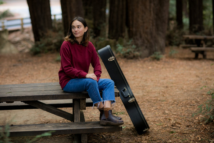 A woman wears a Pladra Women's Crew Neck Sweatshirt in Pomegranate Red, sitting on a picnic table in the forest.