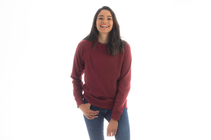 A woman wears a Pladra Women's Crew Neck Sweatshirt in Pomegranate Red and blue jeans, smiling against a white background.