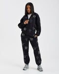 A model wearing a black zip-up hoodie and matching sweatpants set, both featuring a white gothic-style monogram pattern, posed against a light gray studio background.