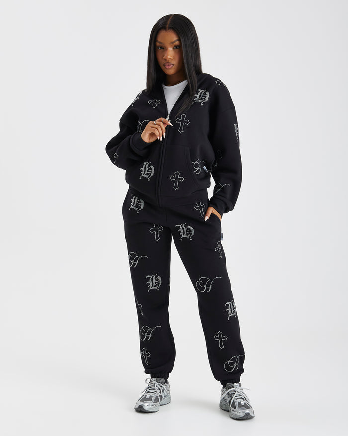 A model wearing a black zip-up hoodie and matching sweatpants set, both featuring a white gothic-style monogram pattern, posed against a light gray studio background.