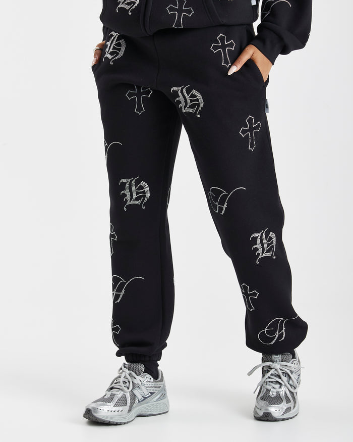 Black sweatpants with an all-over white Gothic monogram print, shown on a model wearing gray and white sneakers against a light gray studio background.
