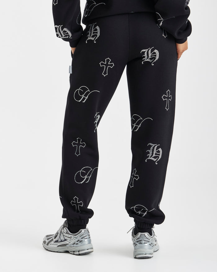 Black sweatpants with an all-over white Gothic monogram and cross pattern, shown on a model from the waist down wearing silver sneakers against a light gray background.