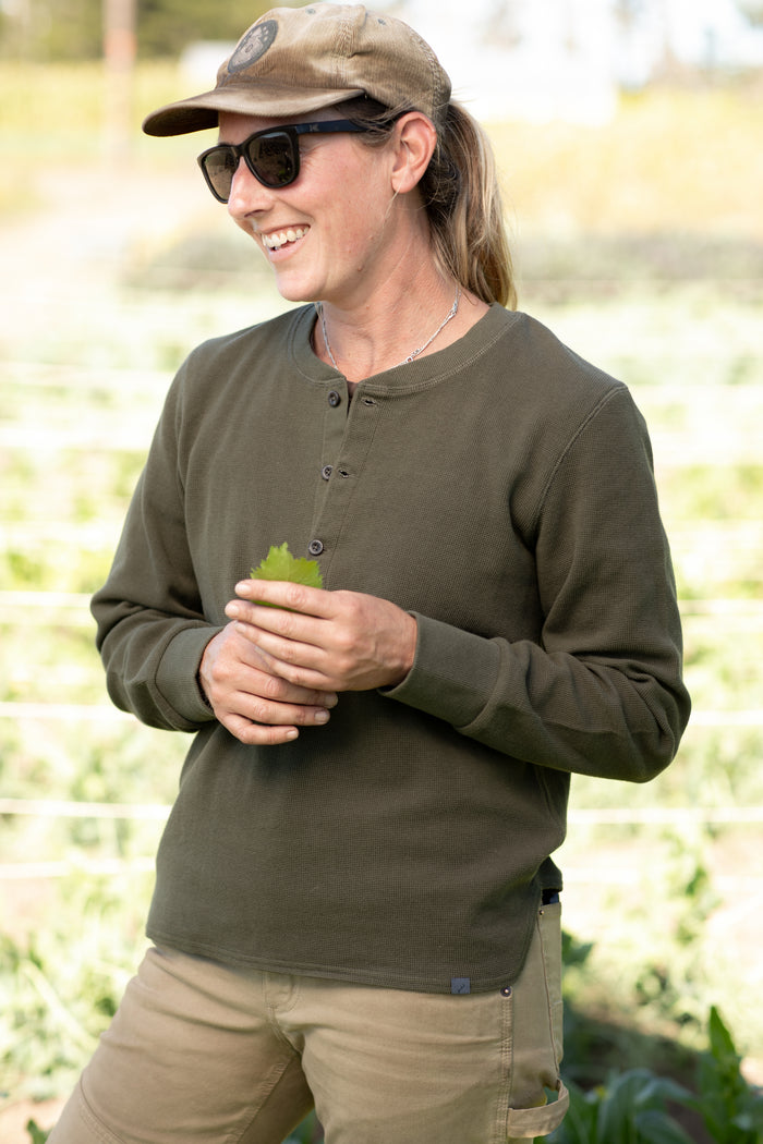 A woman in sunglasses, a khaki cap, and the Women's Elko Henley—Forest Green stands outdoors smiling, holding a green leaf with a blurred field behind her.