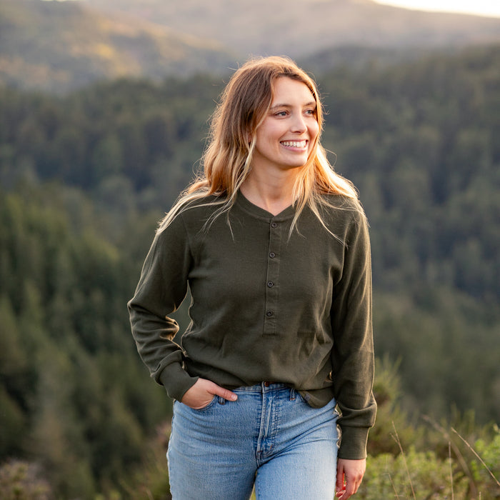 A young woman with long, wavy hair smiles outdoors, wearing the Women's Elko Henley in Forest Green and light blue jeans, with a blurred backdrop of forest and mountains.