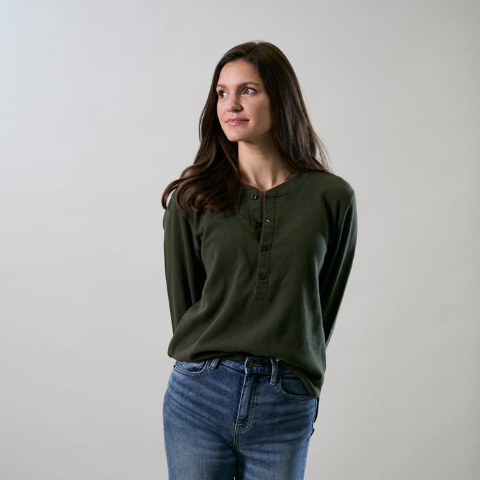 A woman with long brown hair, wearing the Women's Elko Henley in Forest Green and blue jeans, stands against a plain light background, looking slightly to her left with a neutral expression.