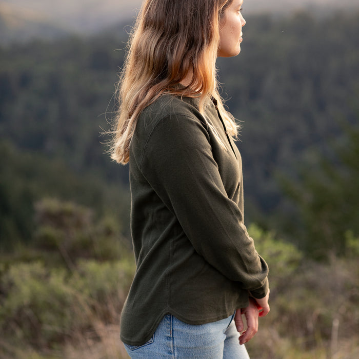 A woman with long, wavy hair wearing the Women's Elko Henley in Forest Green and jeans stands outdoors in profile, with a blurred forested landscape in the background.