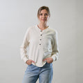 A woman in Pladra's Women's Elko Henley in Oatmeal White and jeans stands with hands in pockets against a light backdrop.
