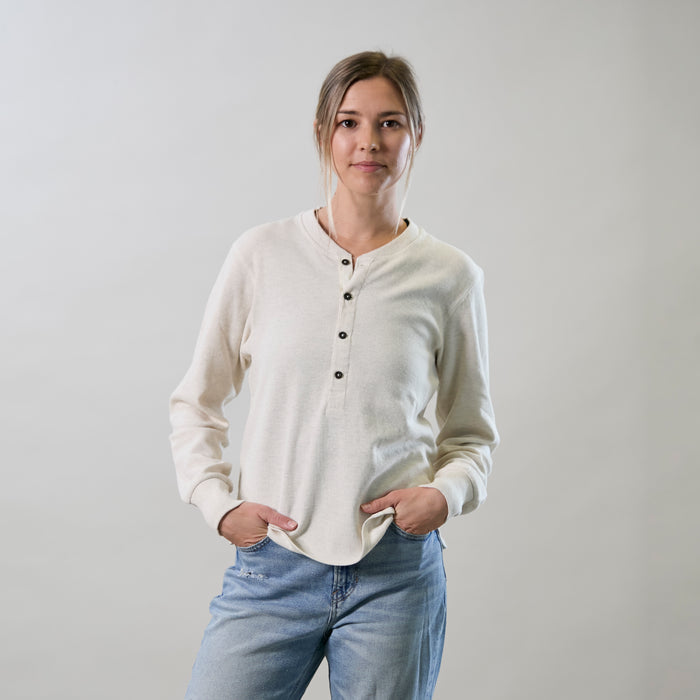 A woman in Pladra's Women's Elko Henley in Oatmeal White and jeans stands with hands in pockets against a light backdrop.