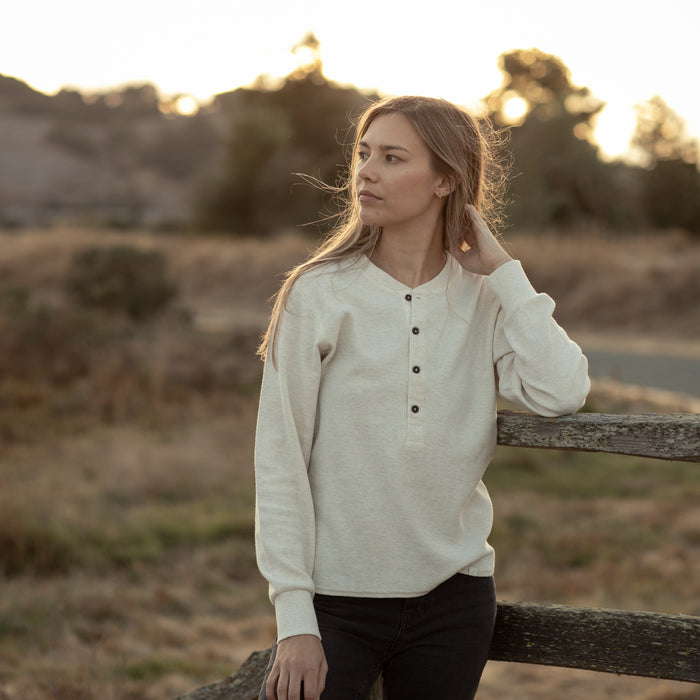 A woman in Pladra’s Women's Elko Henley—Oatmeal White stands by a wooden fence at sunset, gazing to the side.