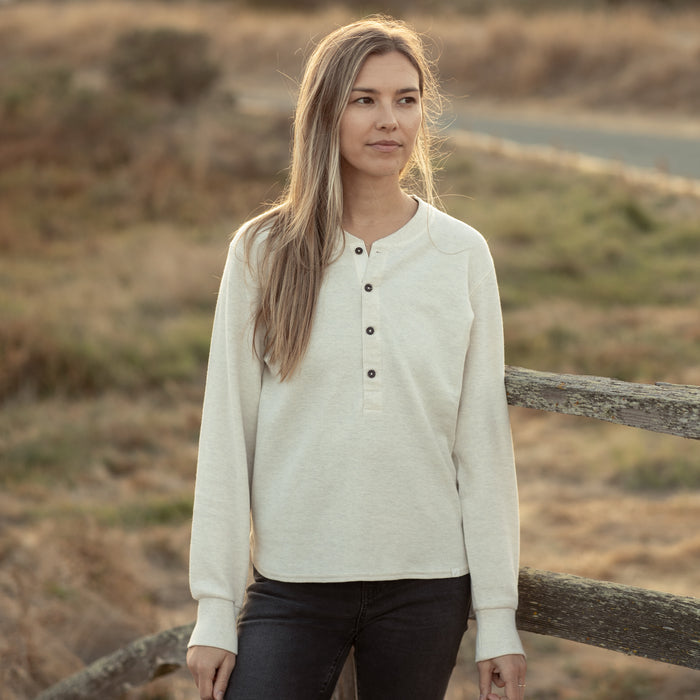 A woman in the Pladra Women's Elko Henley—Oatmeal White stands by a rustic wooden fence outdoors, looking to the side.