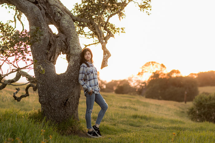 A woman in a Pladra Women's Every Day Double Weave Shirt leans on a tree in a grassy field at sunset.