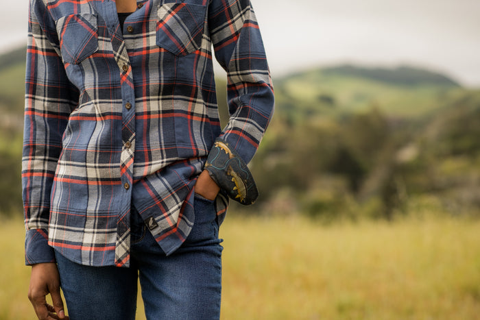 A woman stands in a grassy field wearing the Women's Every Day Double Weave Shirt in Sunset Blue, paired with jeans and a leather bracelet. She has one hand in her pocket; green hills and a cloudy sky blur in the background.