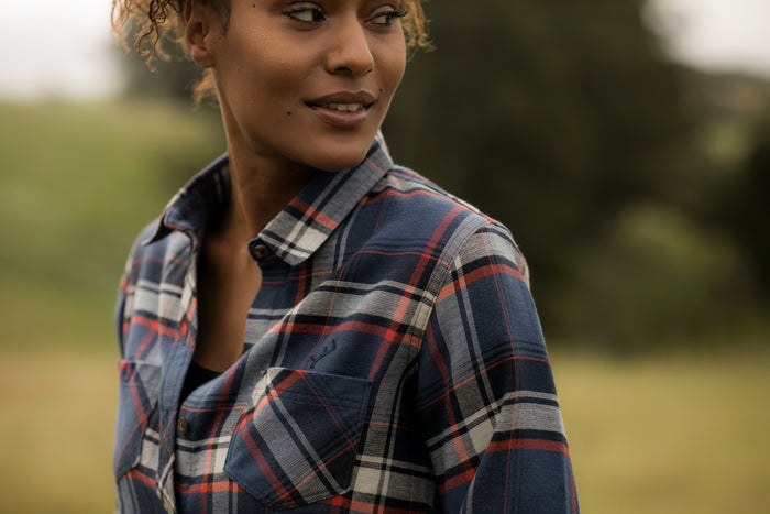 A woman in the Women's Every Day Double Weave Shirt – Sunset Blue stands outdoors, glancing sideways with a gentle smile, as greenery in the blurred background hints at a peaceful, natural setting.