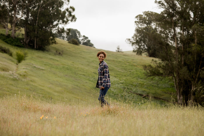A woman in the Women's Every Day Double Weave Shirt—Sunset Blue and jeans smiles in a grassy field, with rolling green hills and tall trees beneath a cloudy sky.