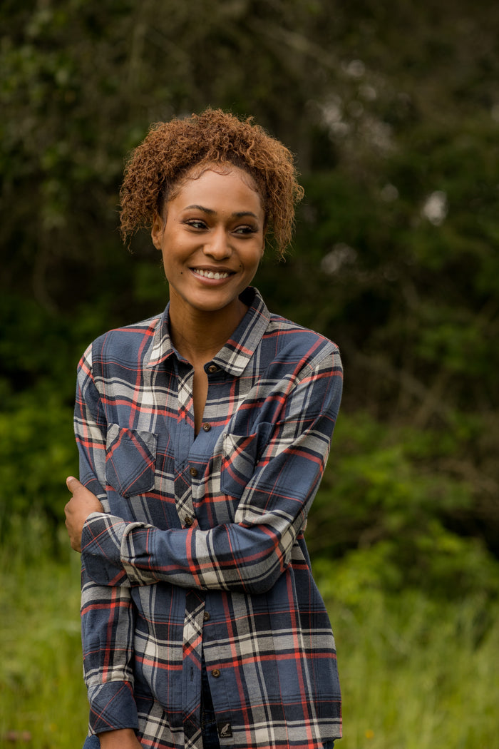 A woman with curly hair stands outside, smiling and looking to the side, wearing the Women's Every Day Double Weave Shirt in Sunset Blue, with blurred greenery and trees in the background.