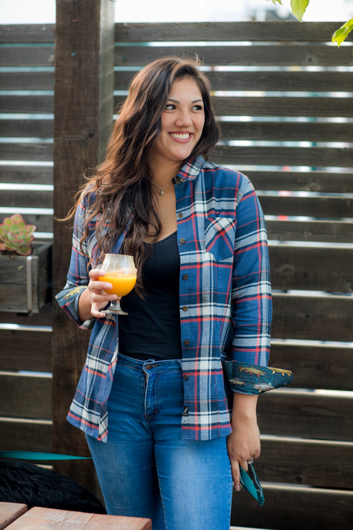 A woman in the Women's Every Day Double Weave Shirt- Sunset Blue and jeans stands outdoors, smiling with a glass of orange juice. Wooden slats and a potted succulent appear in the background.