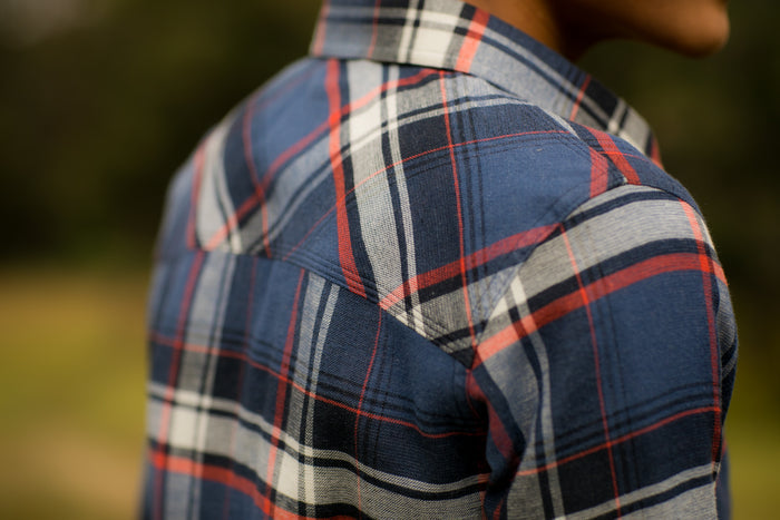 Close-up of a person wearing the Women's Every Day Double Weave Shirt- Sunset Blue outdoors, showing its blue and red double weave flannel pattern and texture. The background is blurred.